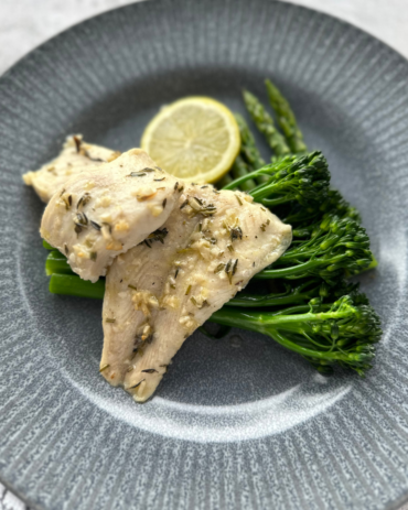 A cooked fillet of lemon and herb sea bass sits atop a bed of green asparagus and broccoli and sliced lemon on a gray plate in an overhead close-up image emphasizing the fresh colors and ingredients of the dish.