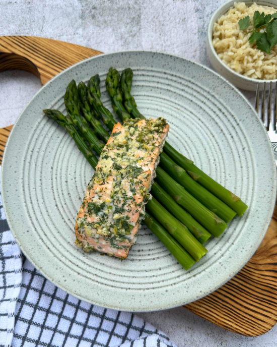 Salmon Fillet with Garlic, Lemon, and Dill, served atop a bed of crisp Asparagus on a light grey plate. The plate rests on an oak chopping board, accompanied by a side bowl of boiled rice adorned with fresh parsley.